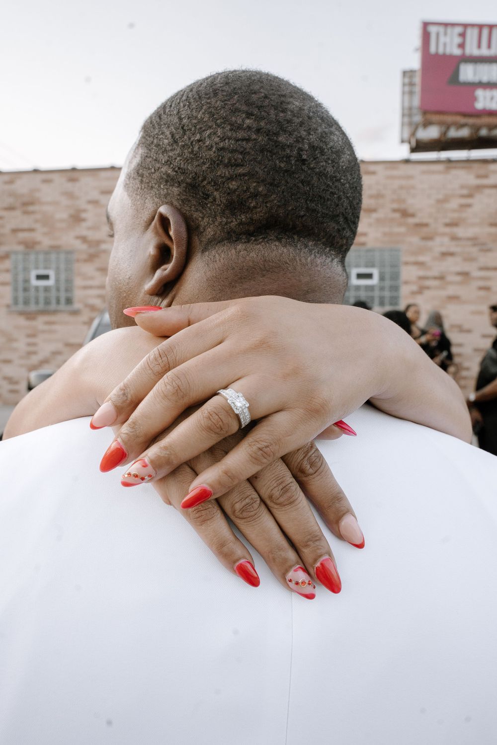 Close-up shot of hands with red nails and an engagement ring resting on a white shirt.