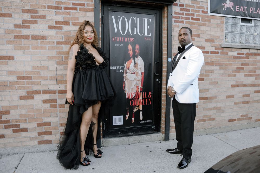 Two people pose in formal attire next to a large Vogue magazine cover display on a brick wall.