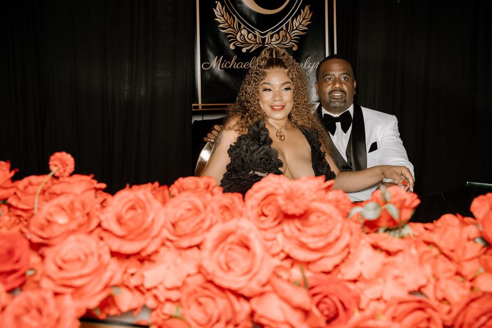A couple sits behind a display of vibrant coral-colored roses at an elegant event.