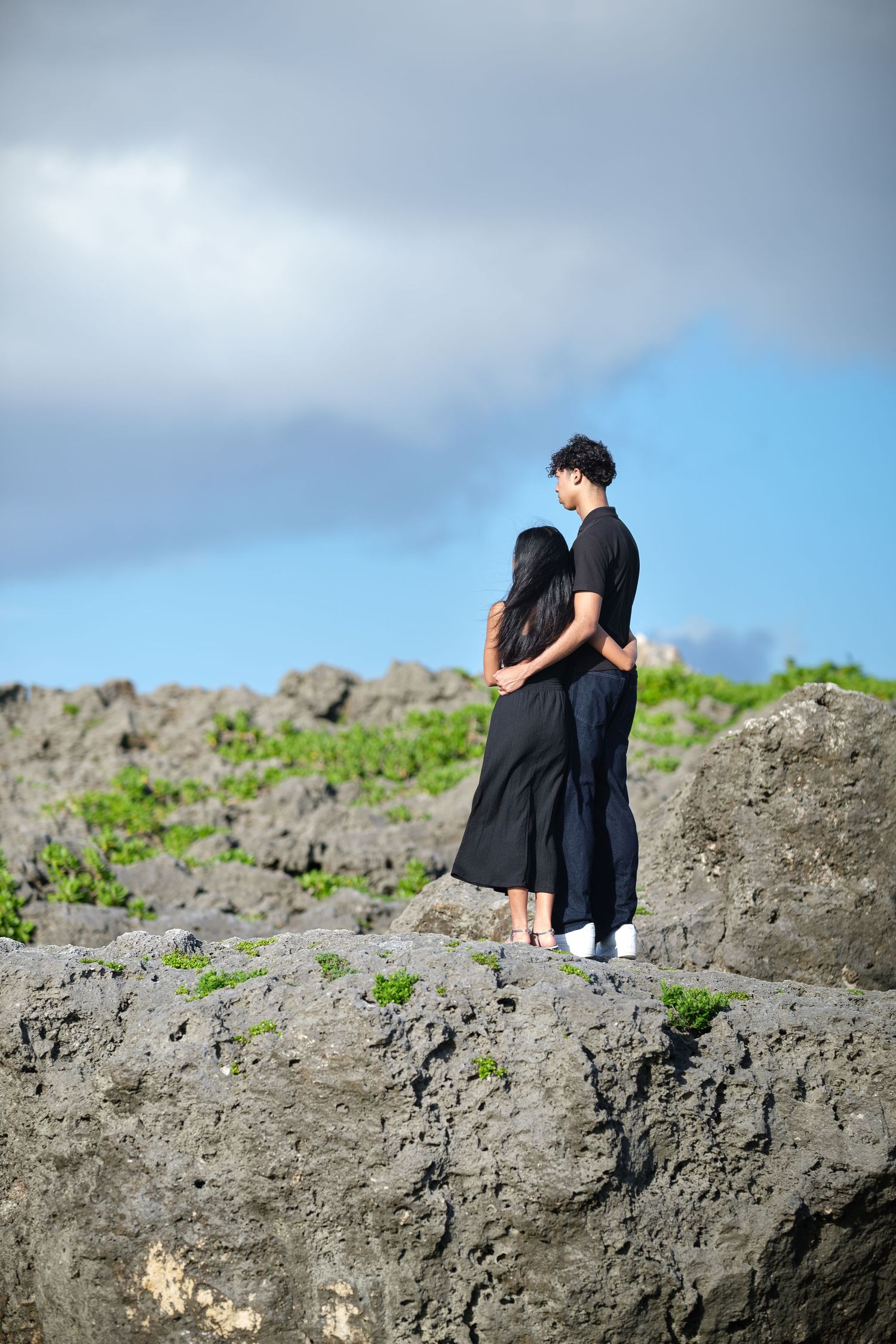 Two people in black outfits sit together on a rocky cliff overlooking a scenic view on a cloudy day.