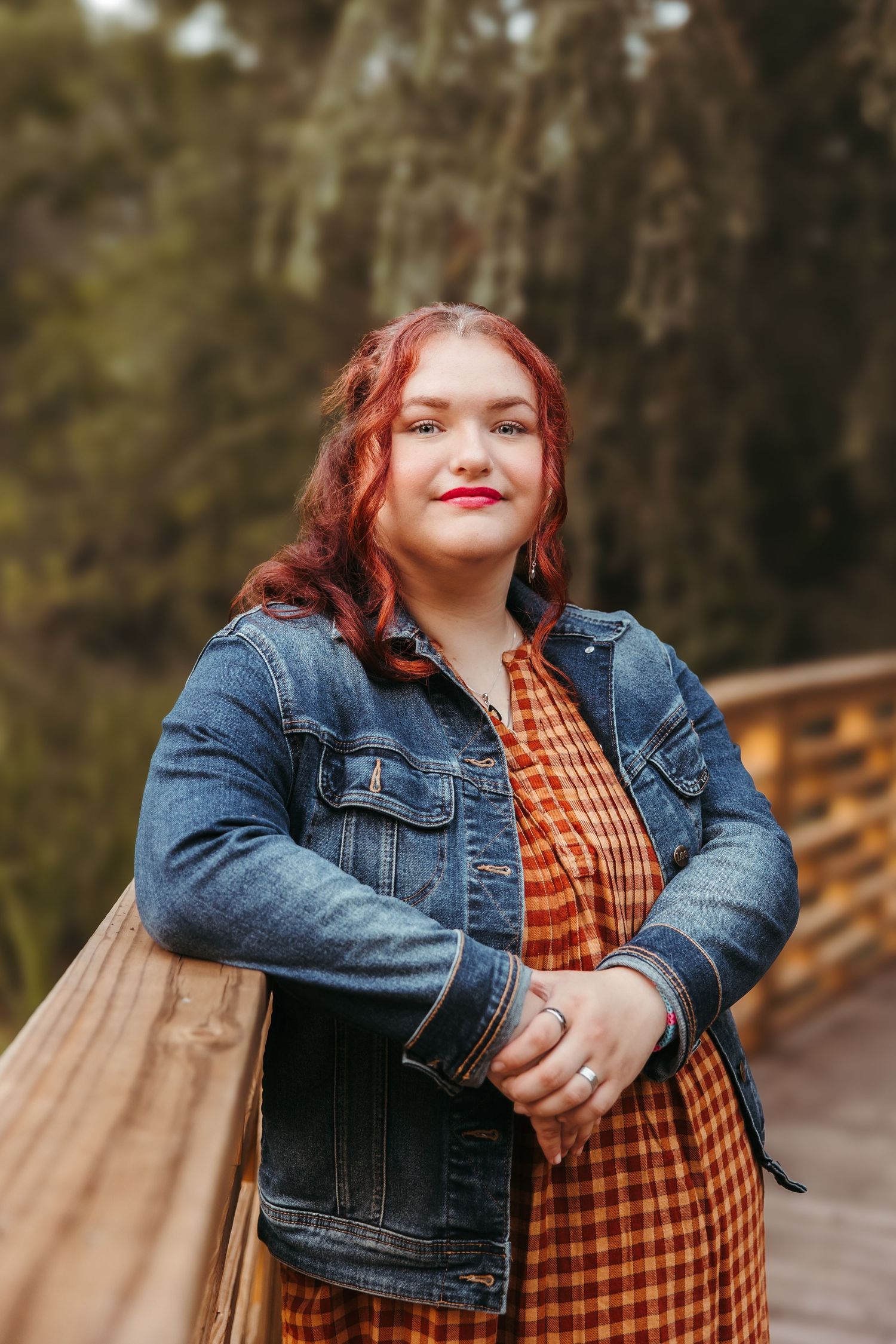 A hs senior in a denim jacket and orange plaid dress stands on a wooden bridge in an autumn outdoor setting. Metro Detroit Senior Portraits