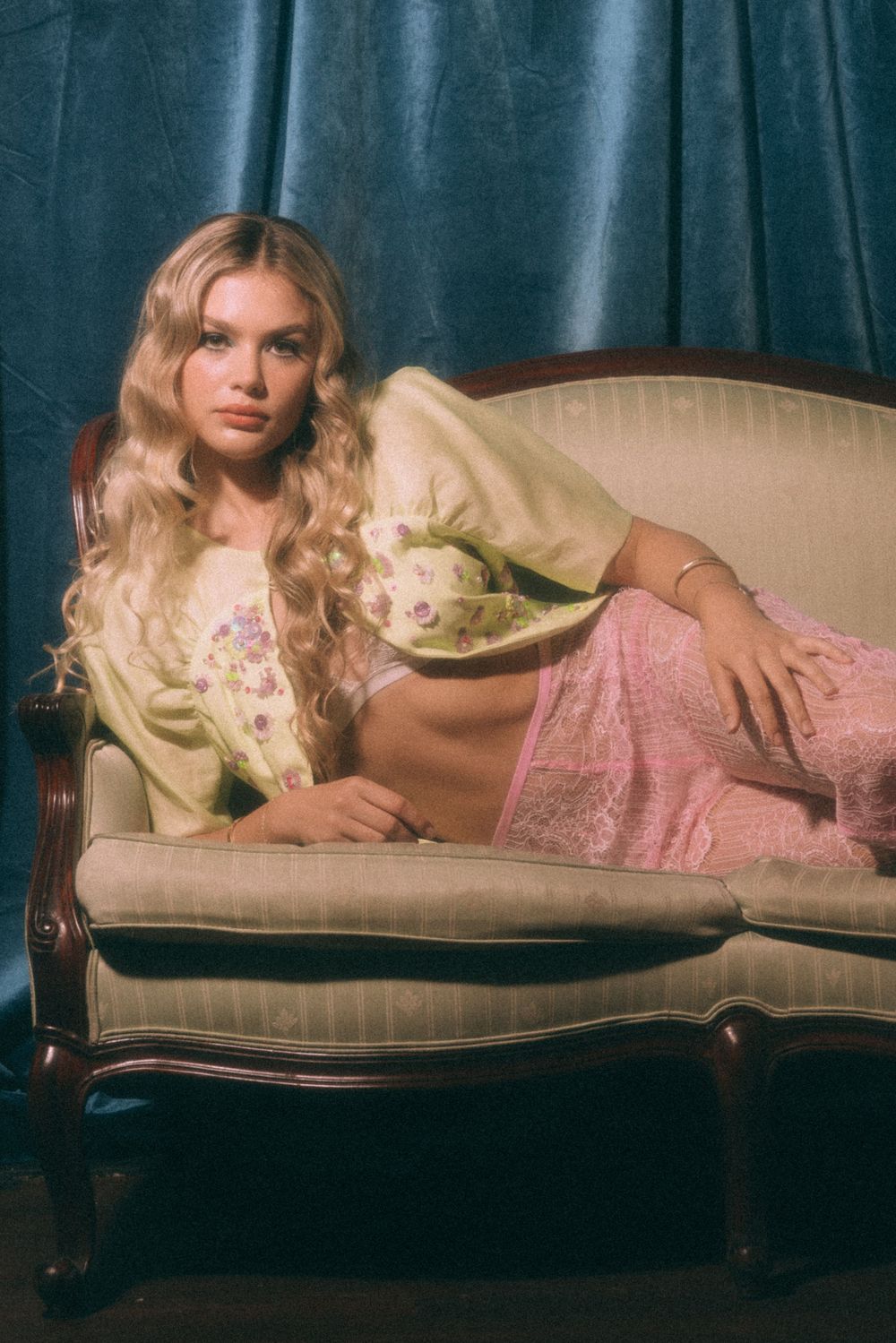 Dreamy boudoir scene with model reclining on antique sofa wearing cream cardigan and pink lace dress.