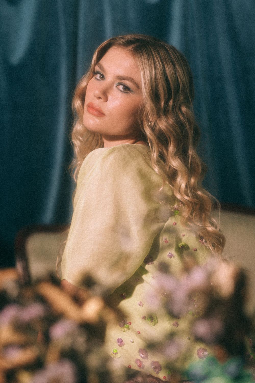 Soft focus portrait with yellow sweater and floral details against dark curtain background.