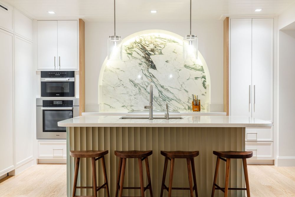 Sleek kitchen featuring curved marble backsplash, white cabinets, and wooden barstools at island.