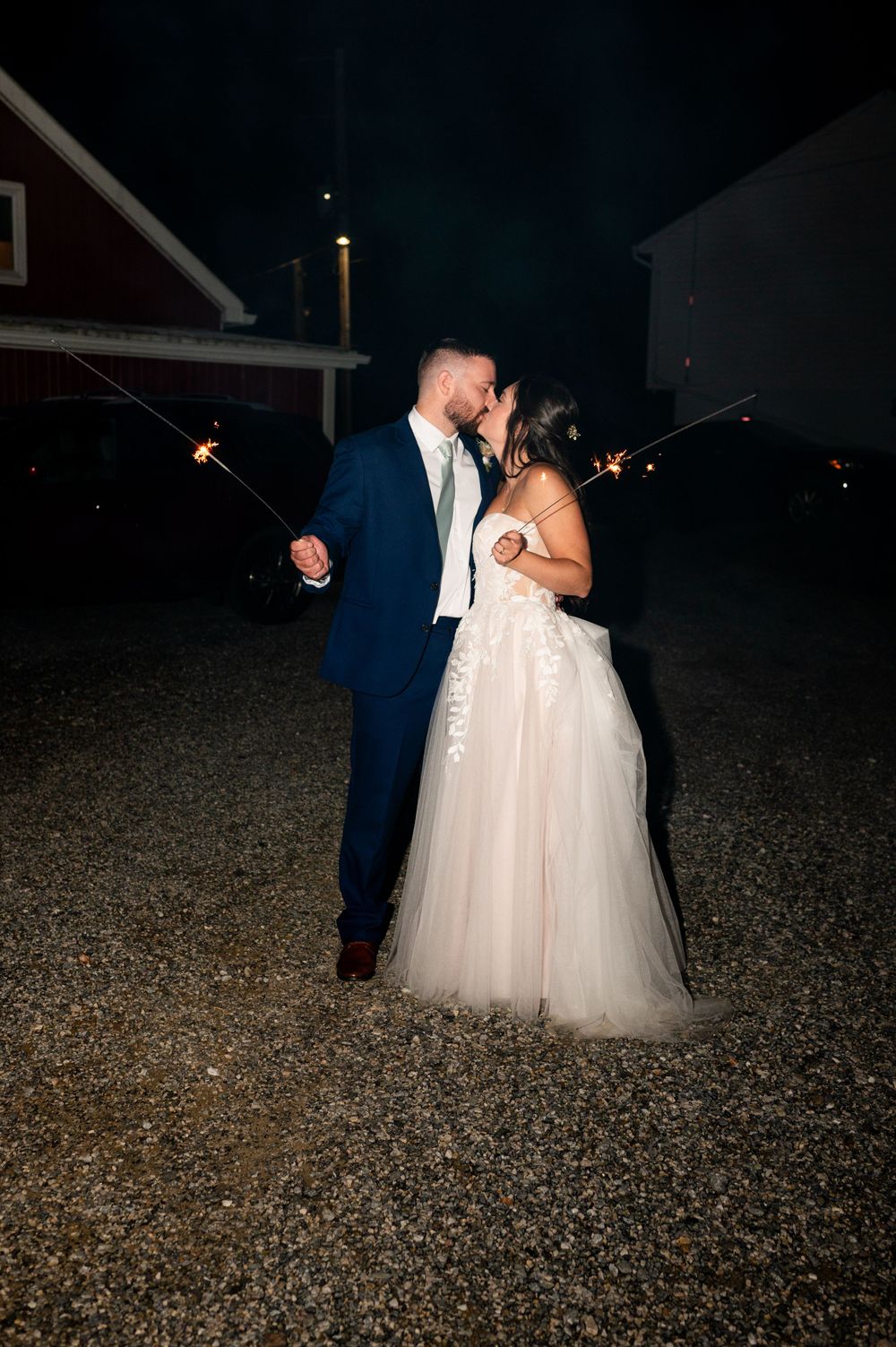 A wedding couple shares a romantic nighttime moment outdoors holding sparklers against a dark background with barns.