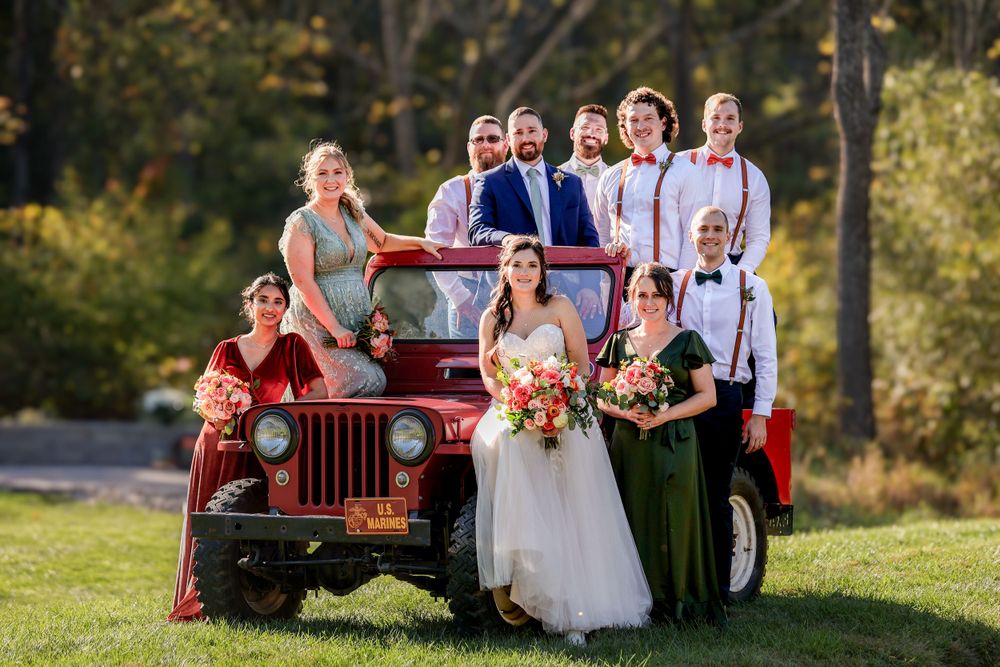 Wedding party poses with vintage red Jeep on a sunny autumn day outdoors.