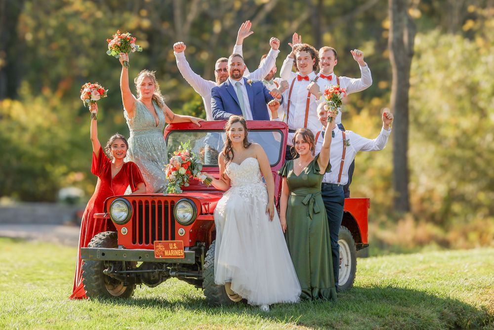 Wedding party celebrates joyfully while posing on and around a vintage red Jeep in an outdoor setting.