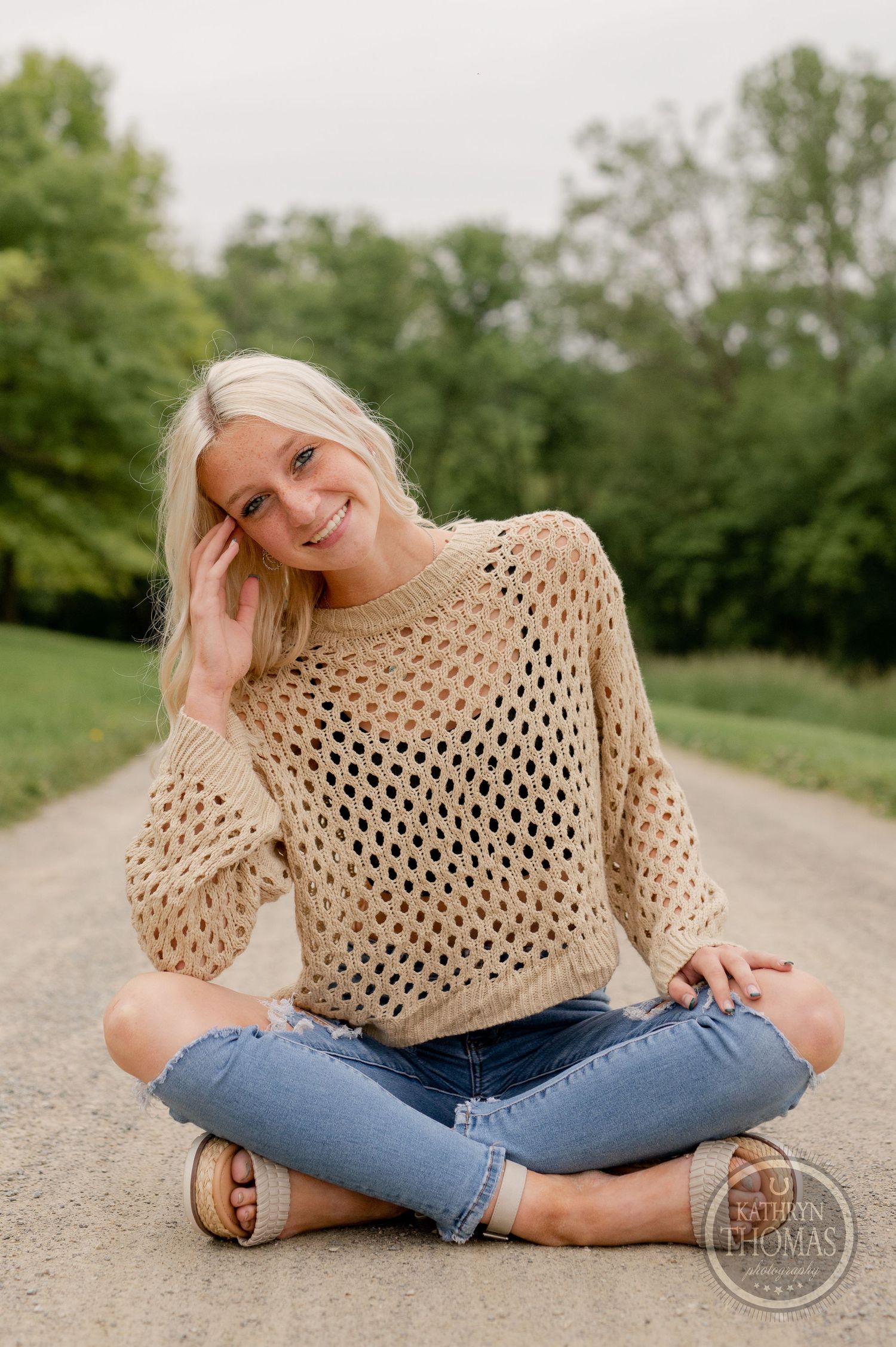 Playful outdoor portrait in crossed-leg pose wearing a beige knit sweater.