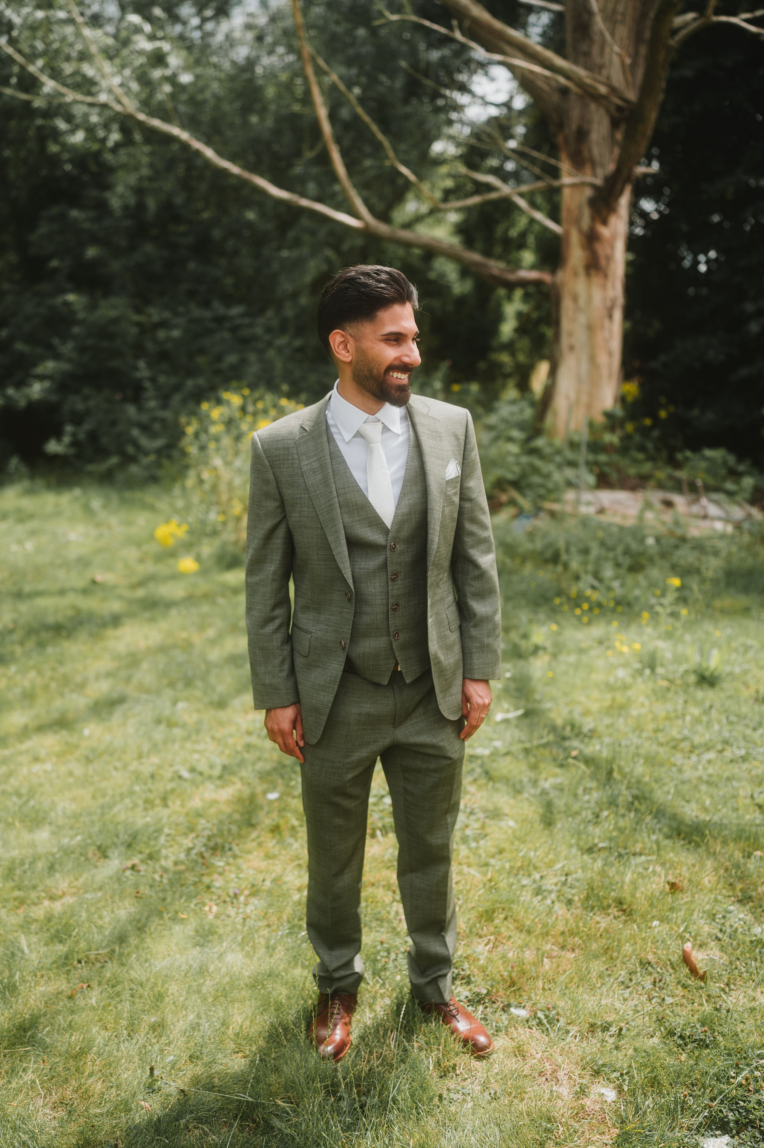Wedding style portrait of a groom in a tailored gray three-piece suit standing outdoors in a garden setting.