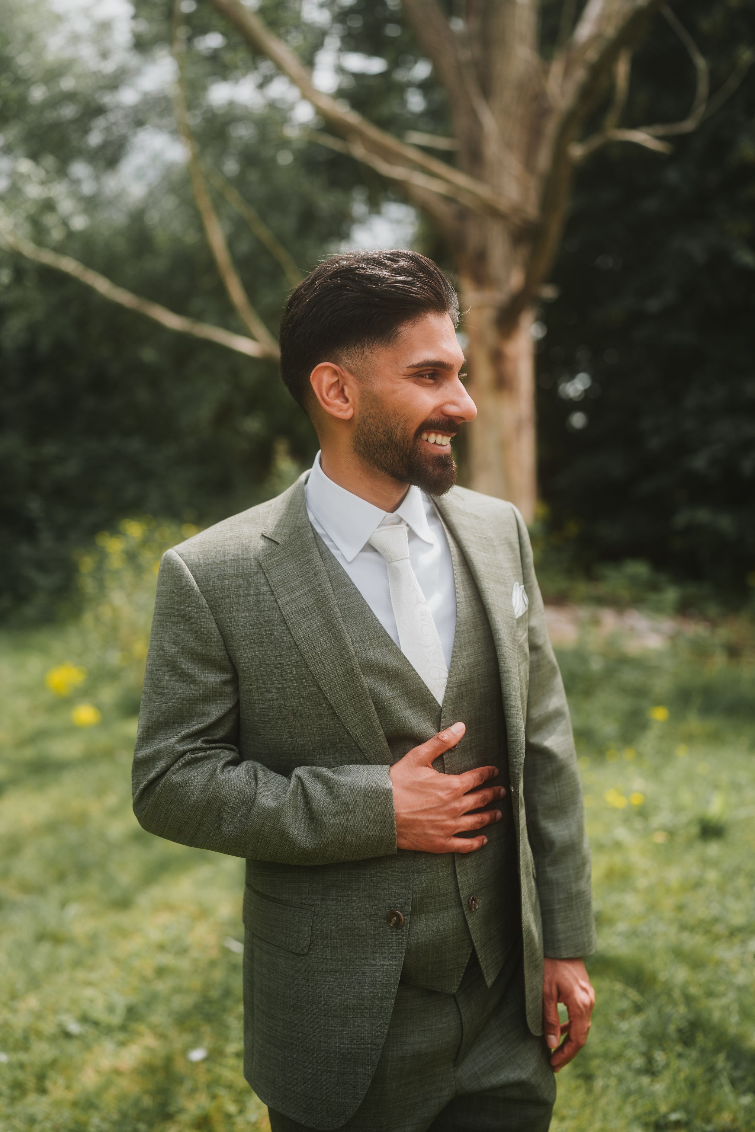 Portrait of a smiling groom in a gray suit adjusting his jacket against a natural outdoor backdrop.