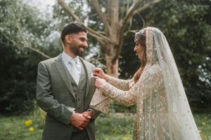 Bride in ornate lace dress with veil shares a tender moment with groom in gray suit during outdoor wedding.