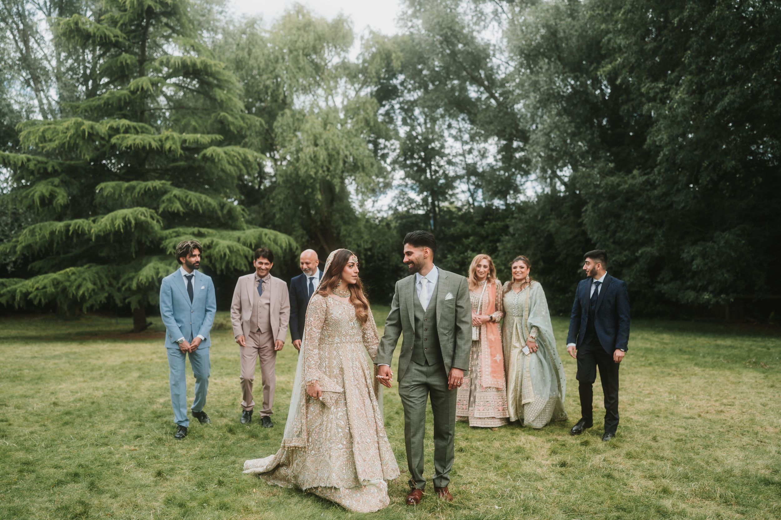 Wedding party poses on lawn with couple in center, bride in beige lace and groom in gray suit.