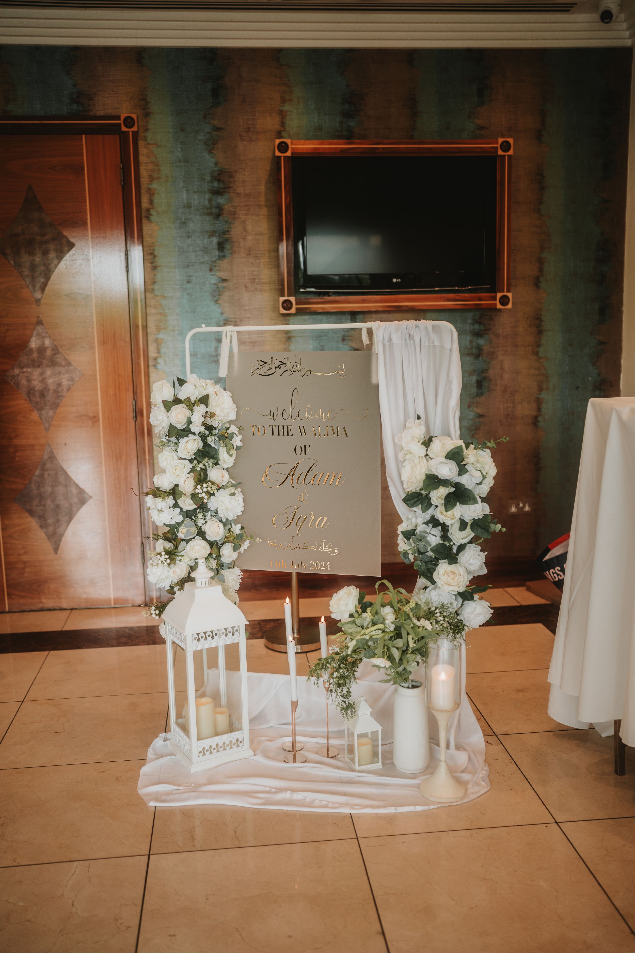 Elegant wedding welcome sign display with white floral arrangements and lanterns against textured wall.