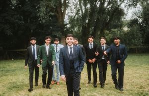 Wedding party of groomsmen in dark suits pose behind the groom in an outdoor garden setting.