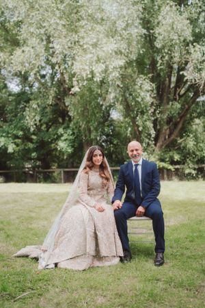 Newlyweds pose in a garden setting with the bride in an ornate beige gown and groom in dark blue suit.