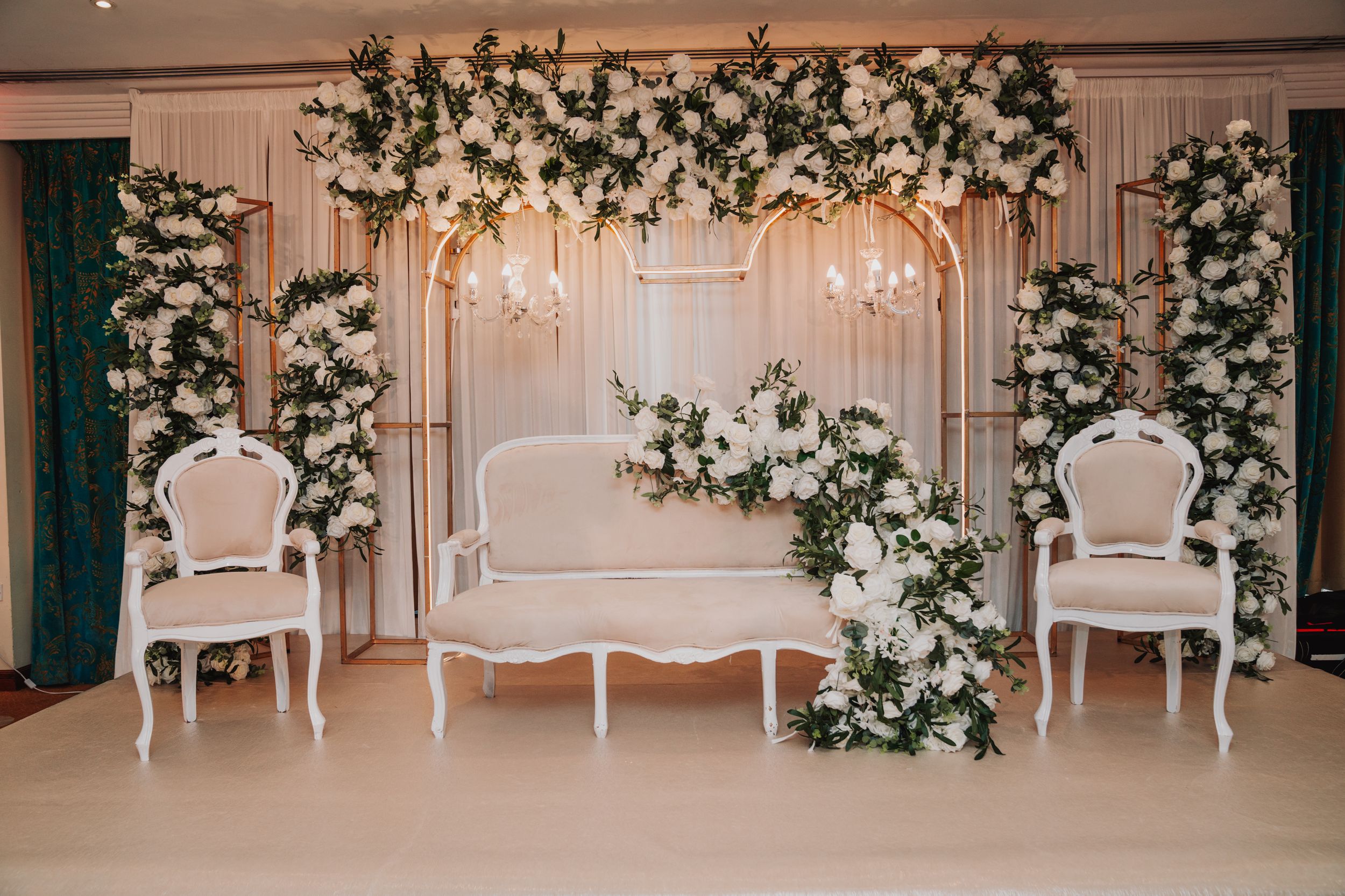 Elegant white wedding stage backdrop with floral arch and vintage chairs.