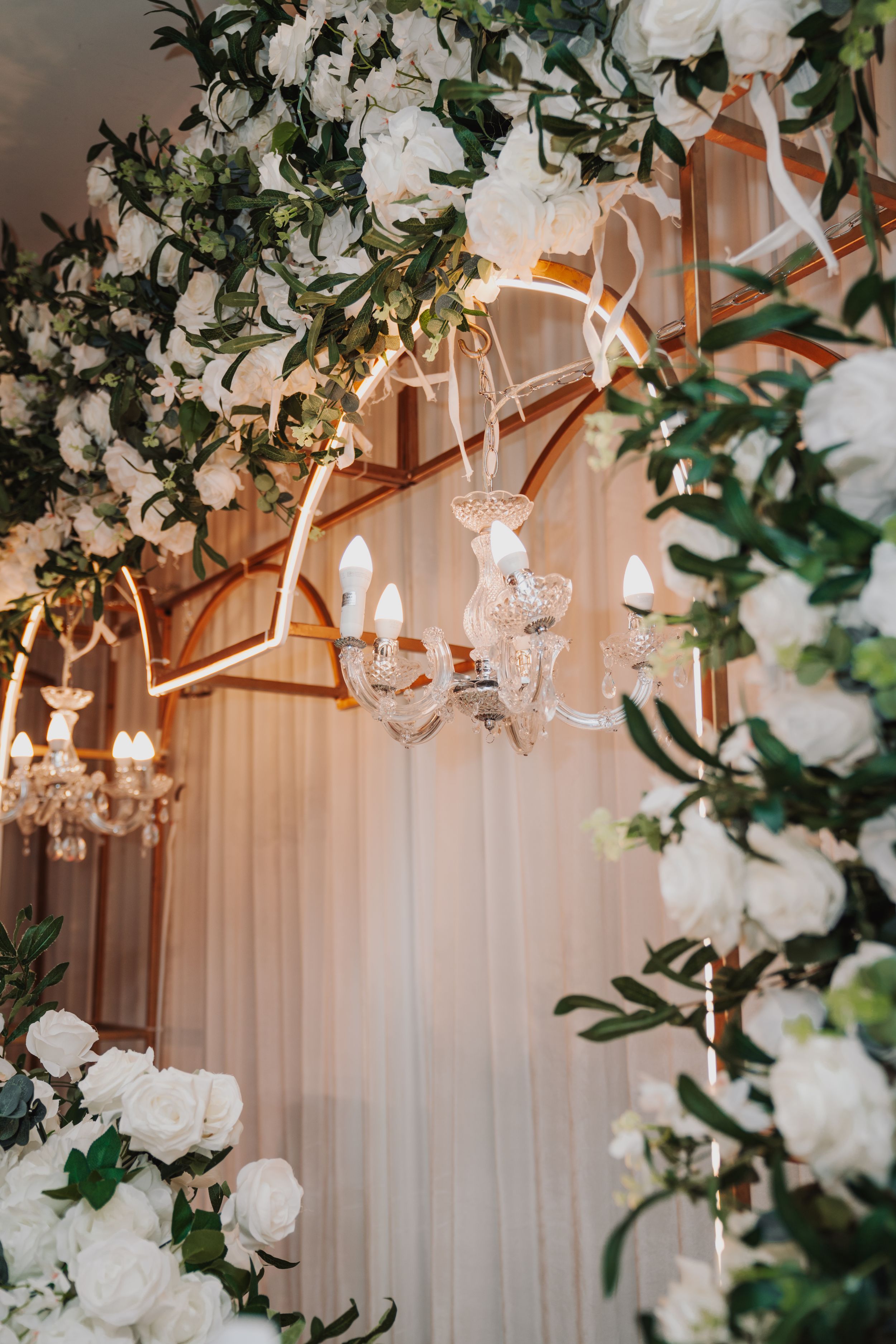 Luxurious wedding decor featuring white roses, crystal chandeliers and greenery against draped fabric.