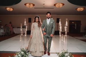 Couple in traditional attire walking between glass pillars with ambient lighting on wedding stage.