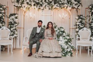 Wedding couple seated on elegant white chairs under floral-decorated arch with hanging crystals.
