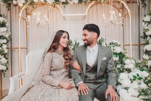 Couple in traditional wedding attire seated in front of floral arch with hanging crystals and soft lighting.