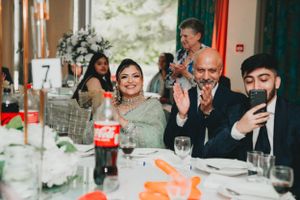 Wedding reception guests at decorated table enjoying festivities with phones and drinks.