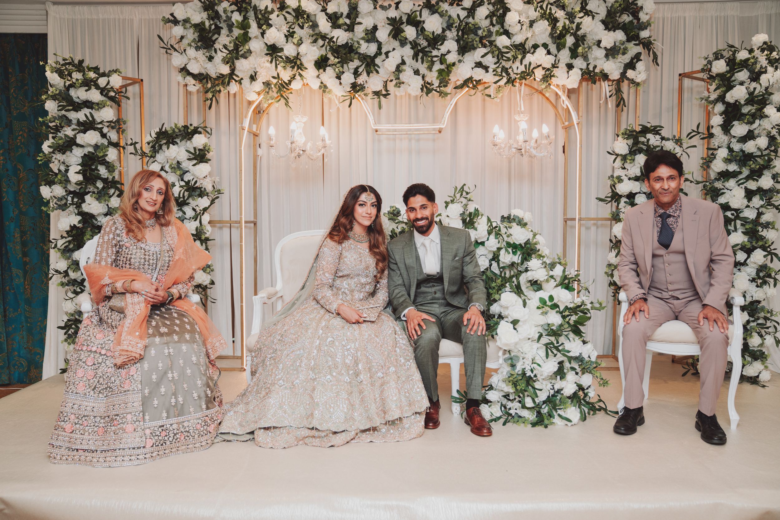Wedding party seated in front of a floral arch backdrop during a South Asian ceremony.