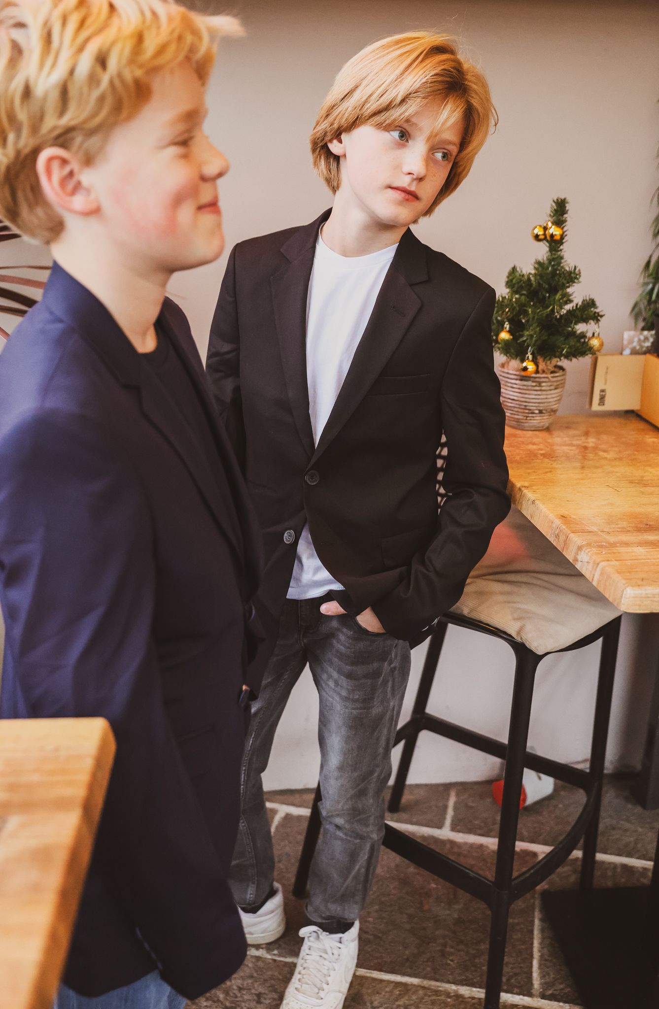 Two casual boys in navy blazers standing near a small Christmas tree indoors.