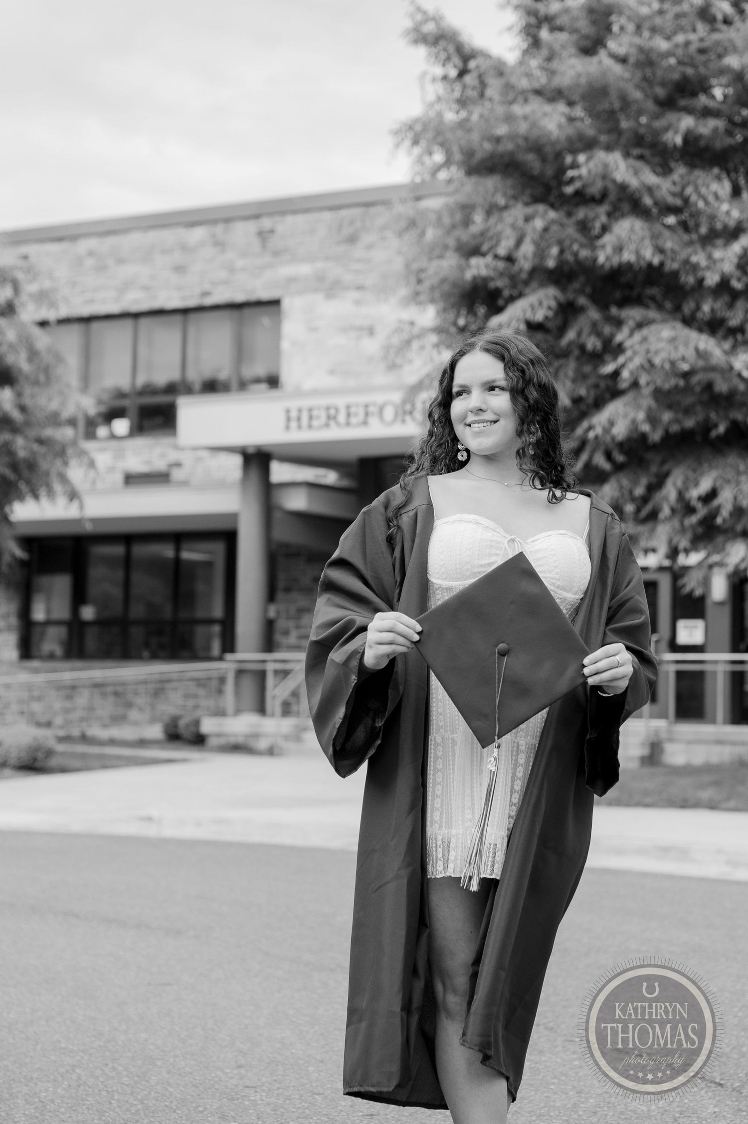 Black and white graduation photo showing graduate holding cap in front of school entrance.