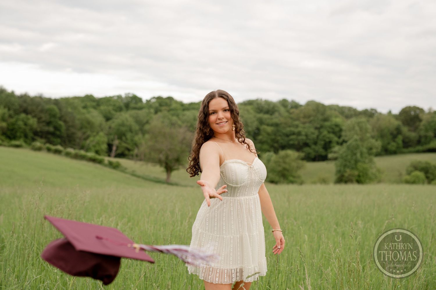 Graduate tosses maroon cap while wearing white dress in rolling green field.