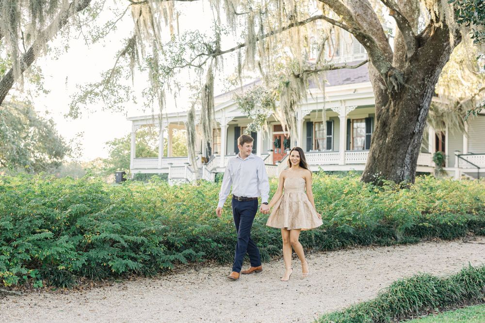 Couple strolling together on path with Spanish moss and historic mansion behind them.
