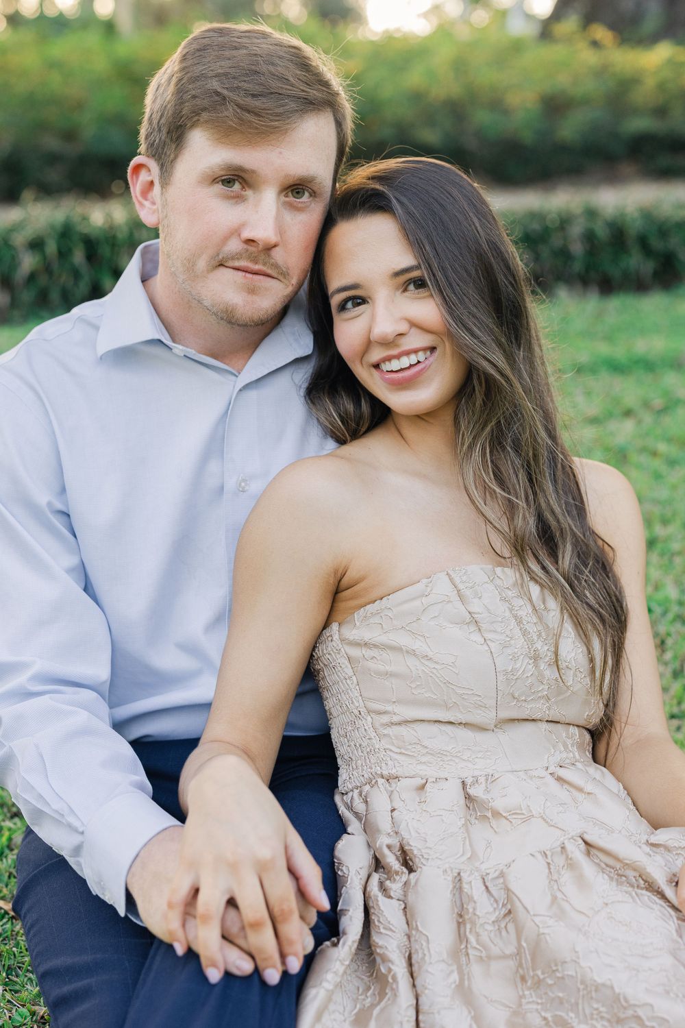 A couple sits together on grass, with her wearing a blush strapless dress and him in a light blue button-down shirt.