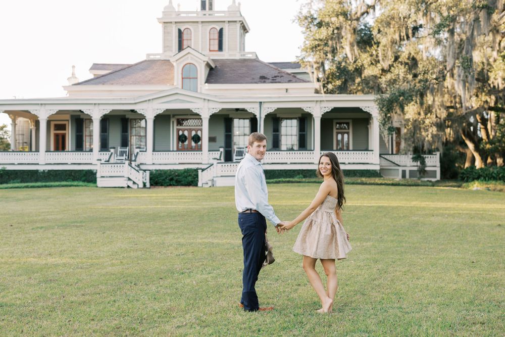 A couple poses playfully in front of a historic white mansion with wrap-around porch.