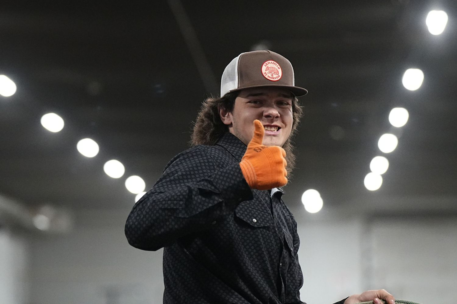 Inside a warehouse with bright lights overhead, someone in a baseball cap and dark jacket holds an orange drink.