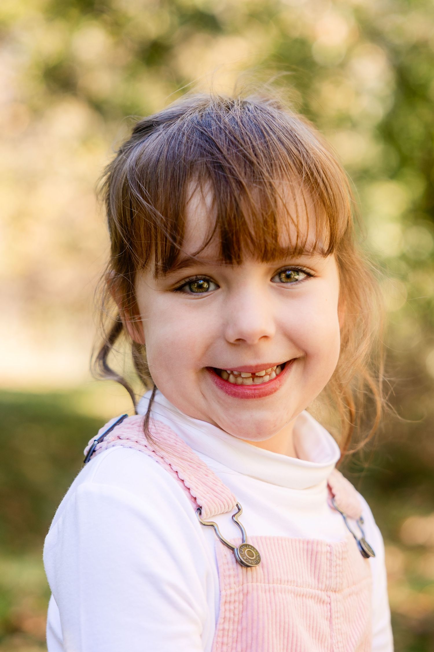 A bright portrait of a child wearing a pink corduroy overall dress and white shirt in an outdoor setting with bokeh background. Novi MI