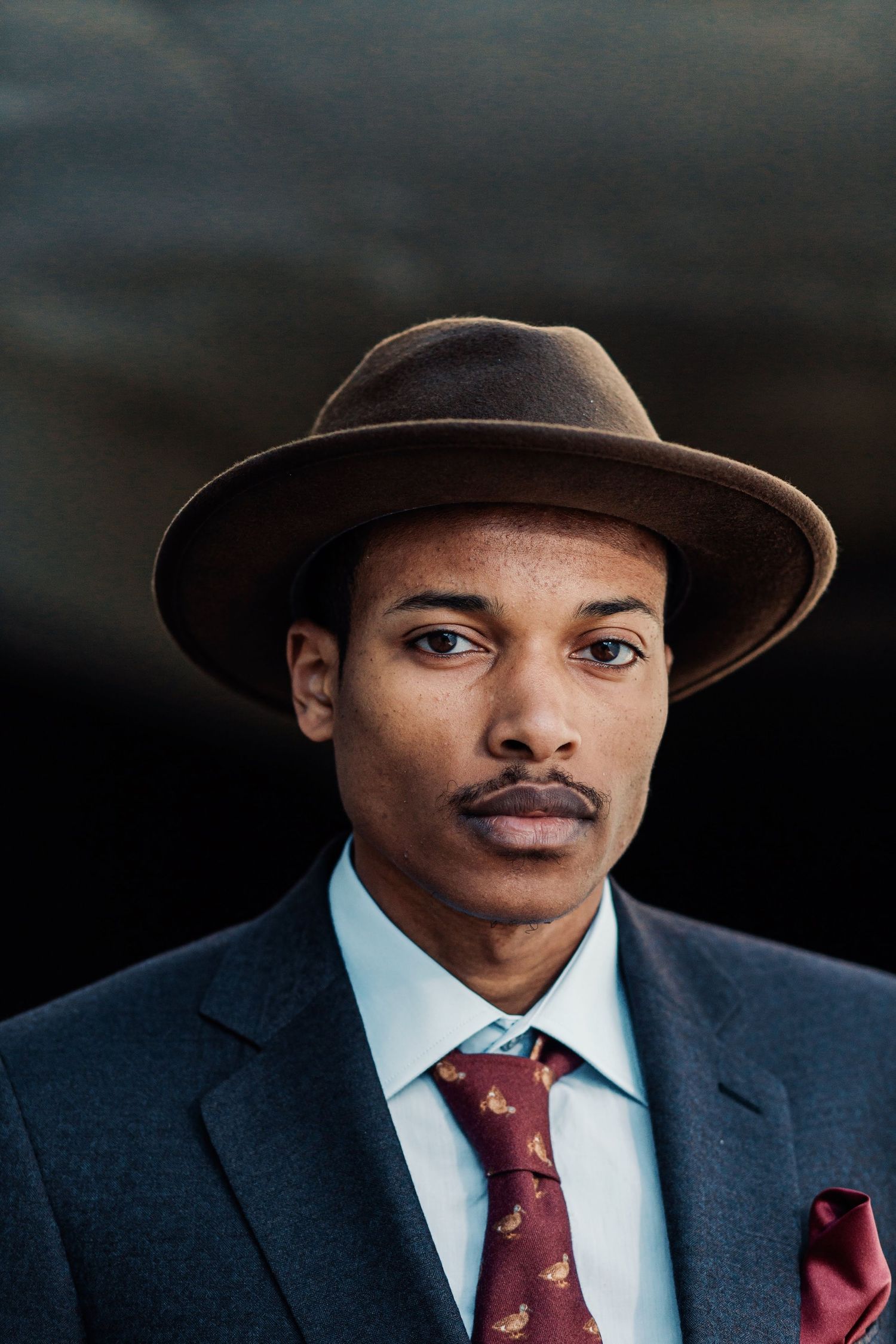 Portrait of well-dressed man in brown fedora hat and navy suit with burgundy tie.