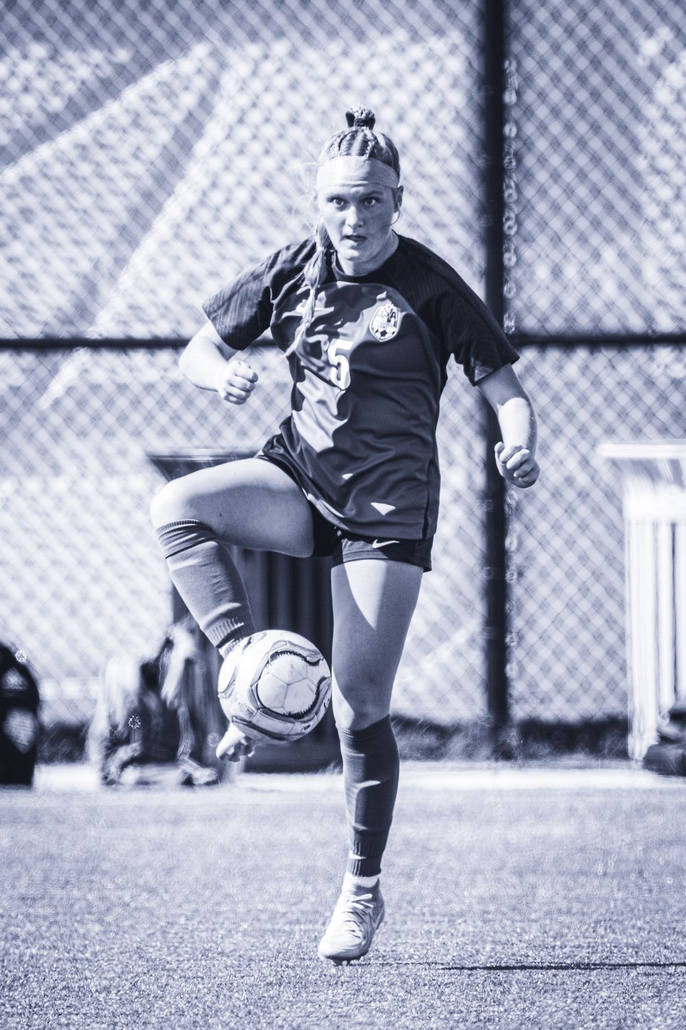 Soccer player dribbles ball during practice against chain-link fence backdrop.