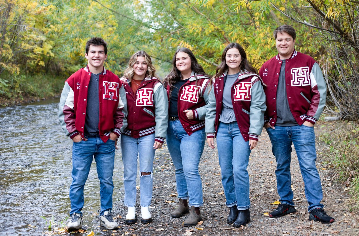 Five people in matching red and gray varsity jackets and jeans stand together on a path near autumn trees.