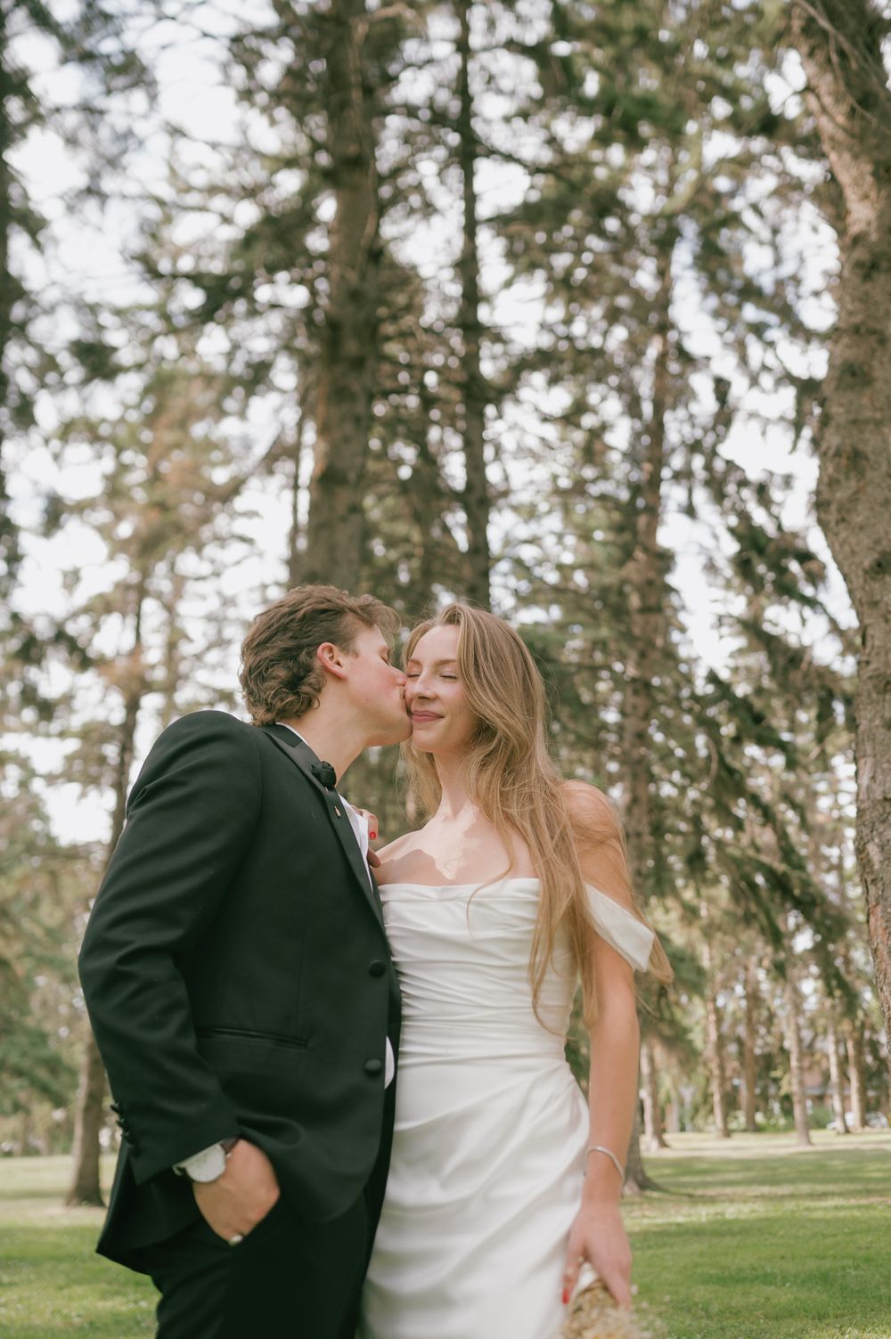Groom in a black suit kisses the cheek of a bride in an off-the-shoulder white satin dress among tall pine trees in a sunny park.