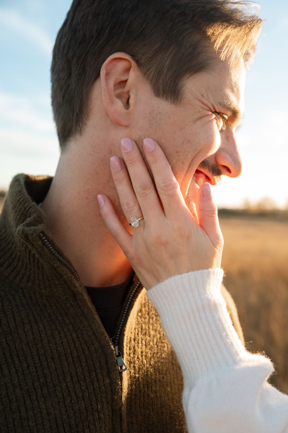 Woman's hand with a diamond engagement ring gently touching a smiling man's cheek in a golden field at sunset.