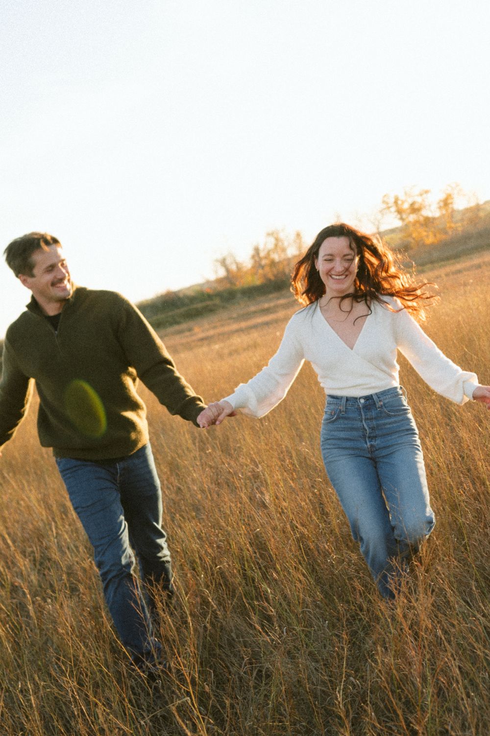 Smiling couple holding hands and walking through a golden grass field at sunset, captured in a candid, joyful engagement-style photo.