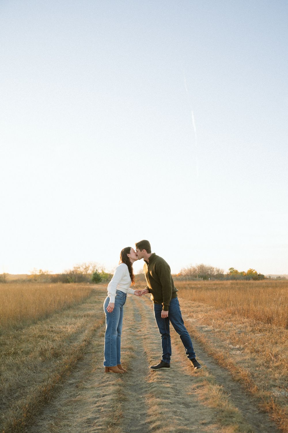 Couple in jeans and sweaters share a kiss on a dirt path through golden autumn grass under a soft, clear sky at sunset.