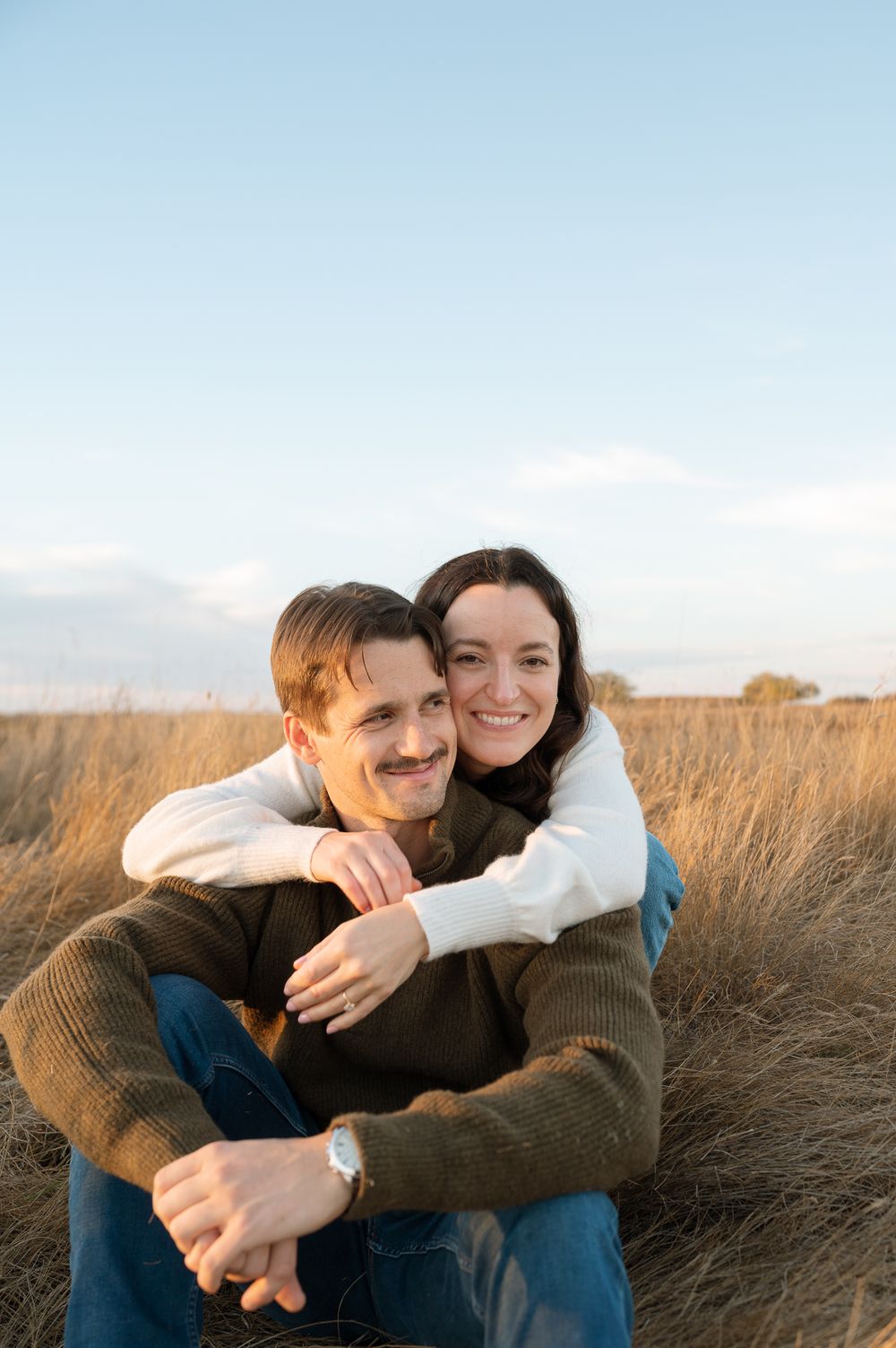 Smiling couple embracing in a golden grass field at sunset, the woman hugging the seated man from behind under a soft blue sky.