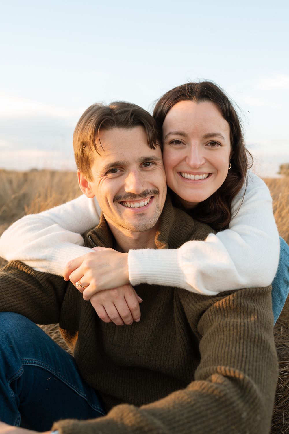 Smiling couple embracing outdoors in a golden grass field at sunset, wearing cozy sweaters, with the woman hugging the man from behind.