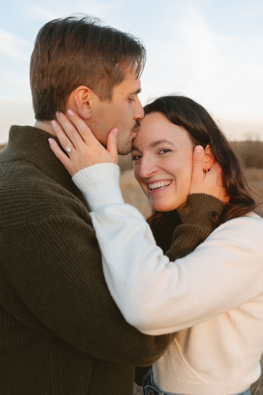 A smiling woman wearing an engagement ring holds her partner's face as he kisses her forehead during an outdoor engagement photo session.