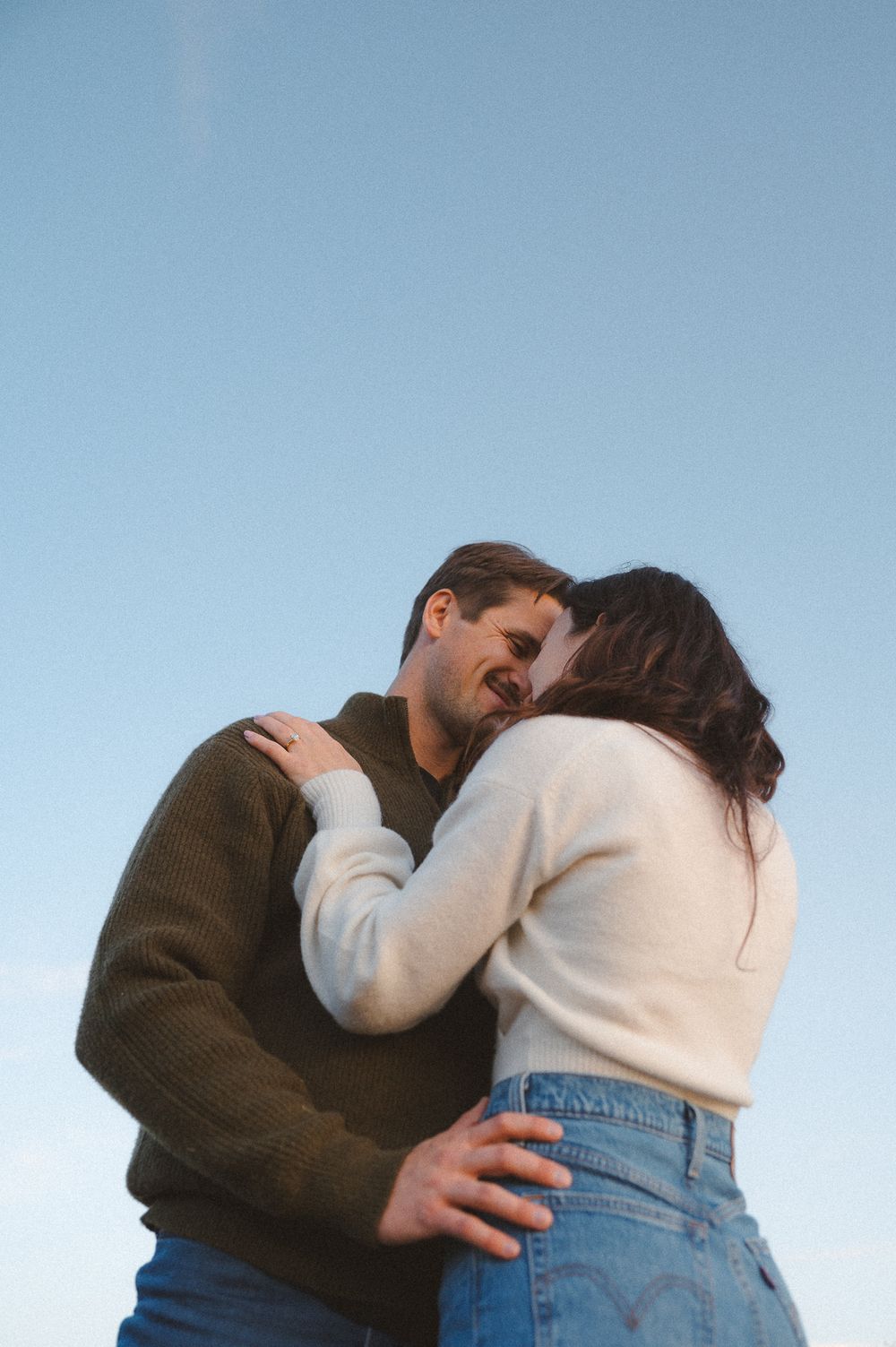 Smiling couple in cozy sweaters embracing against a clear blue sky, a romantic engagement or couples photoshoot moment.