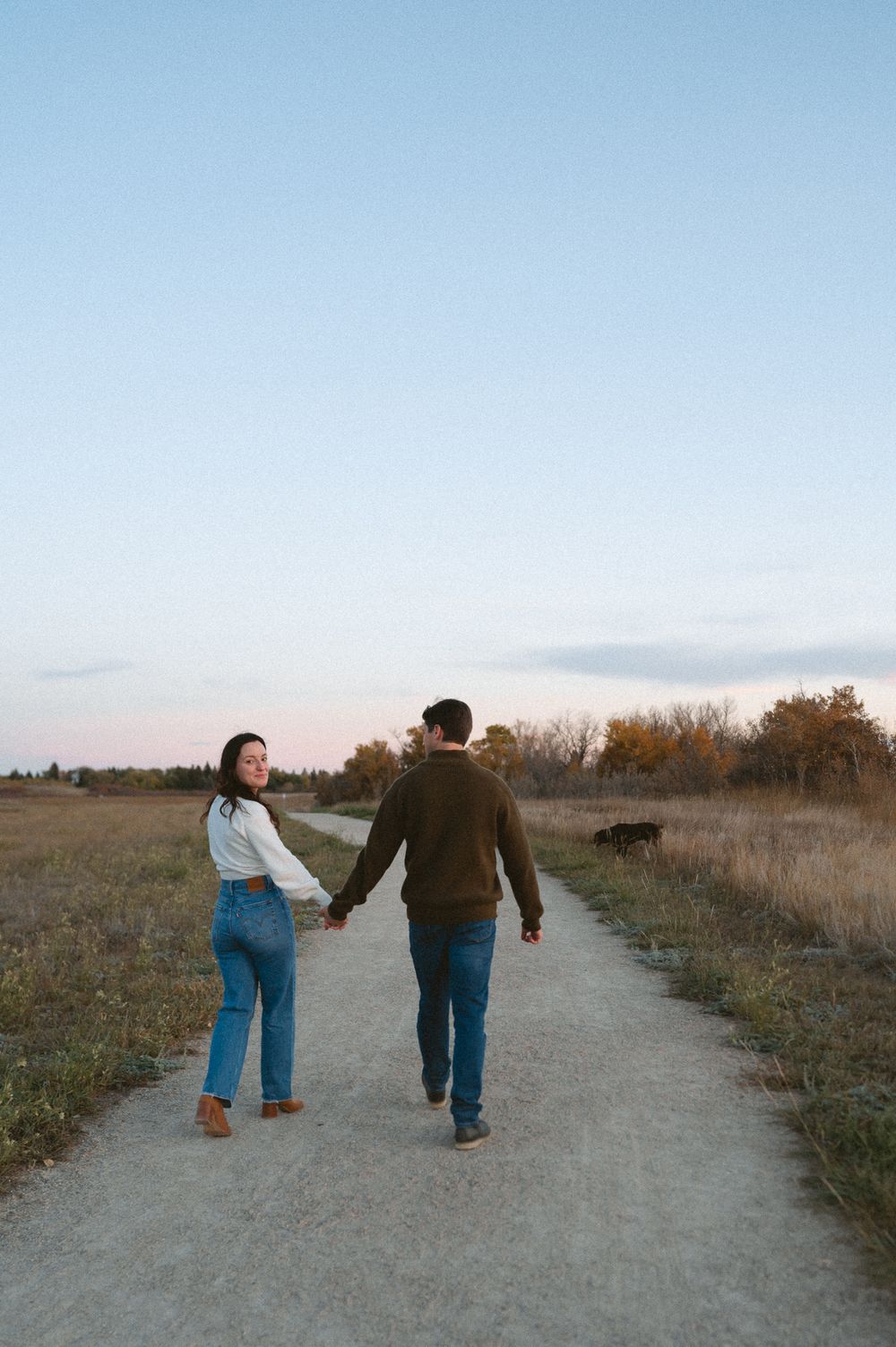 Couple holding hands walk down a gravel path through an autumn prairie at sunset, the woman glancing back at the camera.
