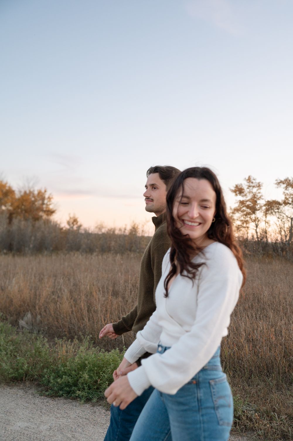 Smiling woman in a white sweater and jeans walks hand in hand with a man along a dirt path through golden autumn grass at sunset.