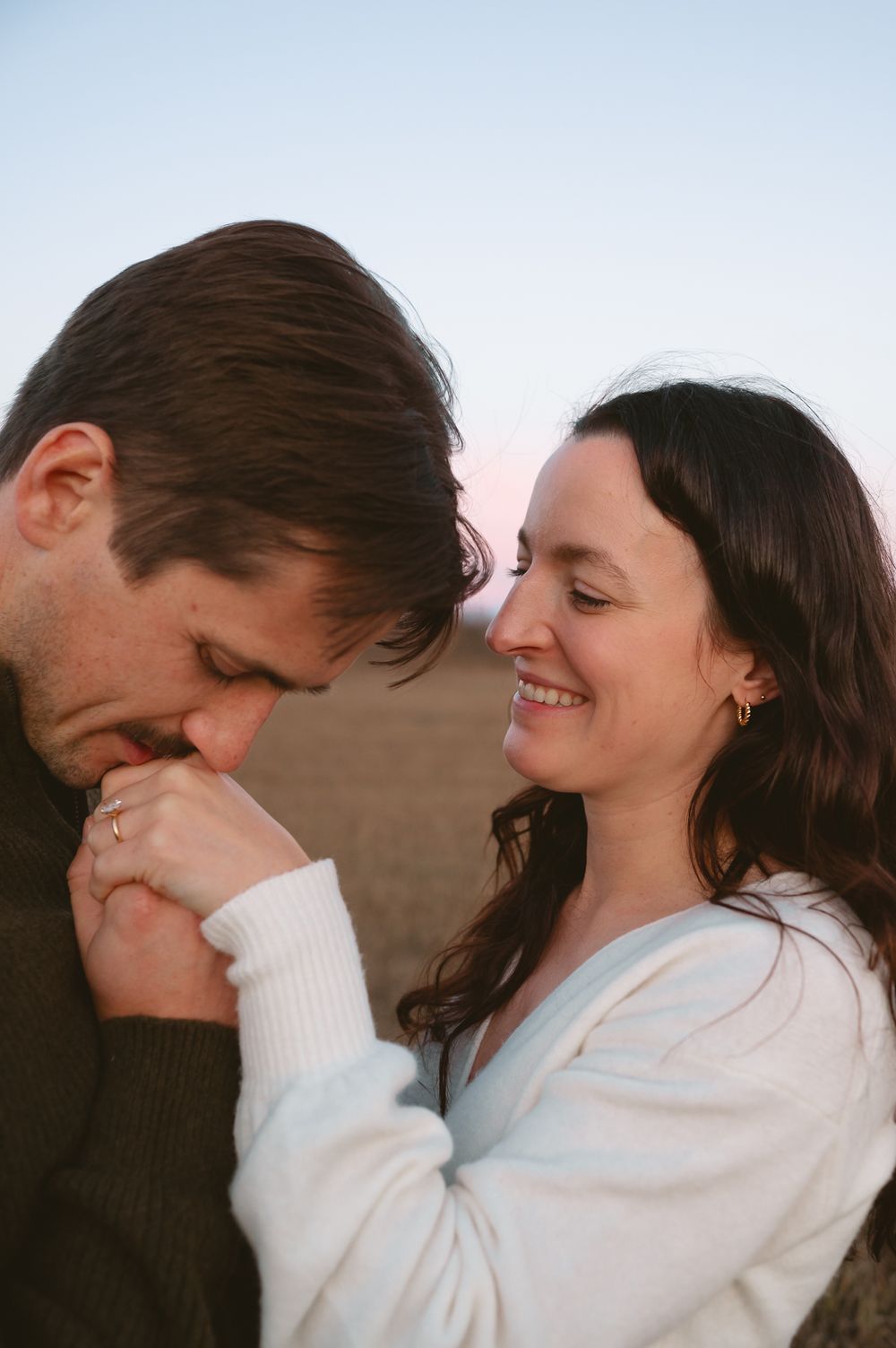 A smiling woman in a white cardigan holds a man's hand as he kisses it tenderly during an outdoor couple's portrait at sunset.