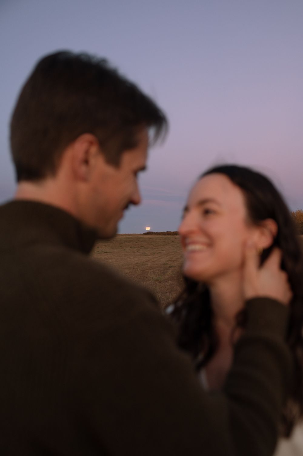 A smiling couple shares a tender moment outdoors at dusk, the woman's hand touching her hair, with a soft purple sky over an open field.