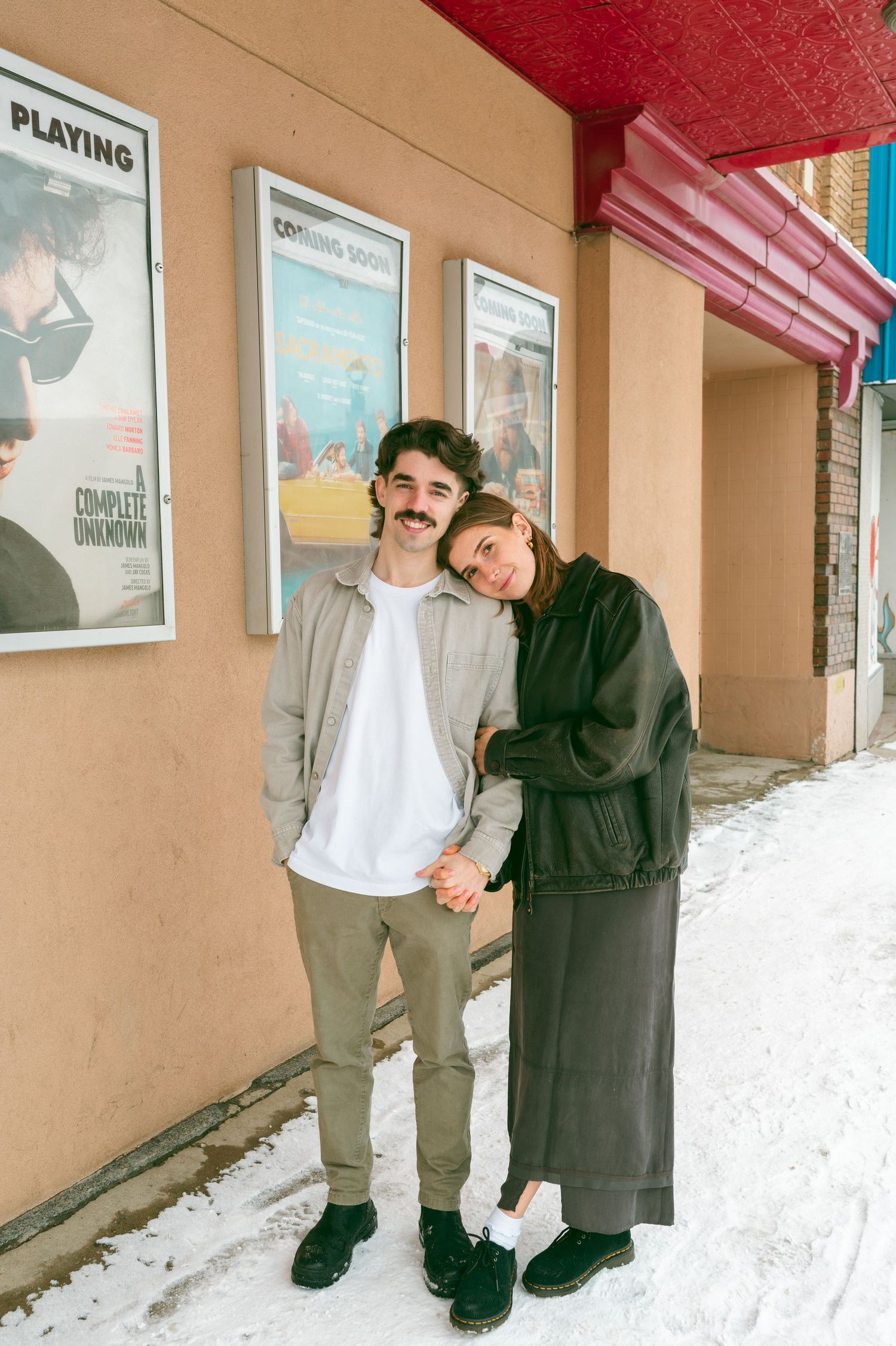 Couple stands together in winter outside an old movie theater with film posters displayed on beige wall.